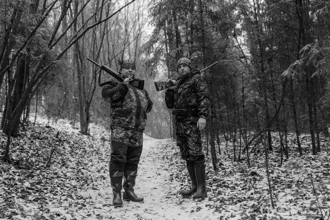 Two hunters in winter camouflage pose with rifles in a snowy forest.
